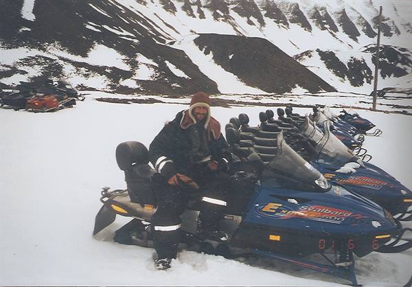 Smiling man in snowsuit beside parked blue snowmobiles on white snow