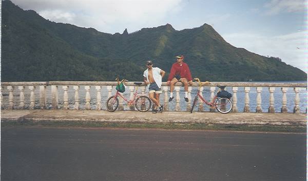Two cyclists rest on a stone balustrade in front of green mountains