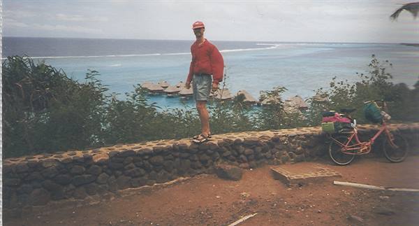 Cyclist in red shirt on stone wall, turquoise lagoon and thatched bungalows, Bora Bora