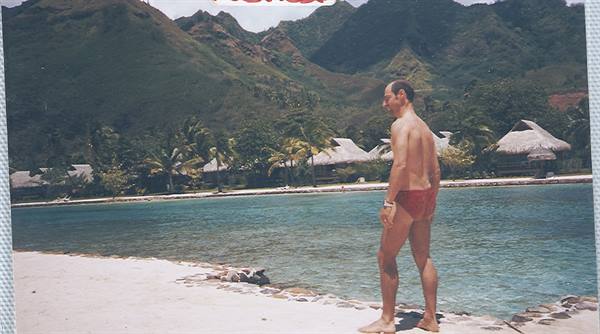 Man in red swimsuit on beach with thatched bungalows and green mountains behind