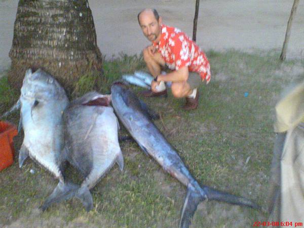 man crouching beside large freshly caught fish under a palm tree