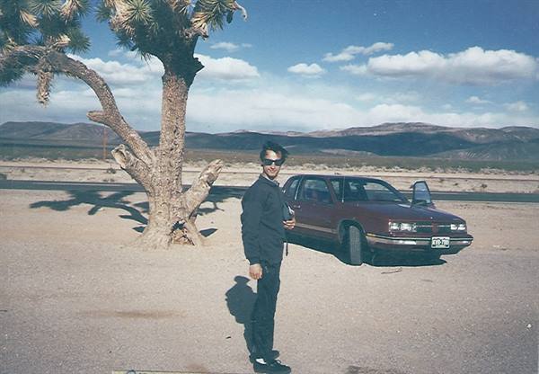 Man in sunglasses beside a red car under a Joshua tree in the desert