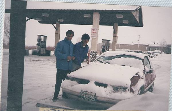 Two young men beside a snow-covered car in front of a gas station