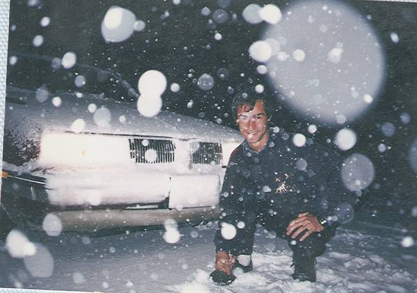 Man crouching beside a snow-covered car at night, snow falling