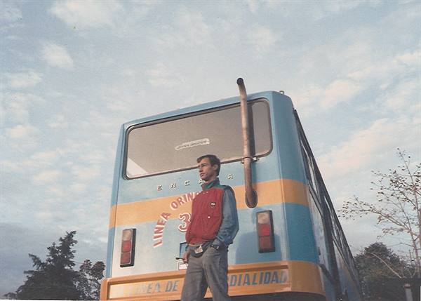 Young man in red jacket standing before an old blue and yellow bus under a cloudy sky
