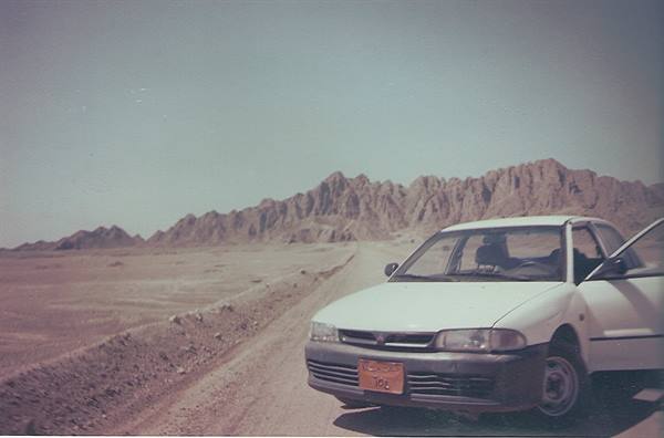 White car parked on desert dirt road, door open, rocky mountains in background