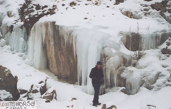 Man beneath frozen waterfall with icicles, Whitehorse sign, snow and rocks