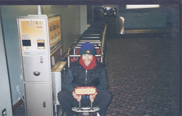 Man in blue cap sitting on airport luggage cart beside a vending machine
