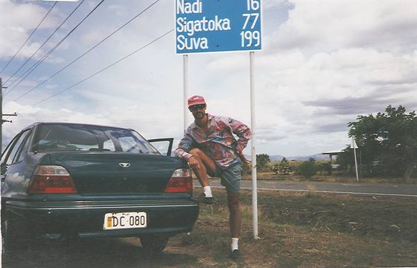 Traveler leaning on a car by a Fiji road sign showing distances