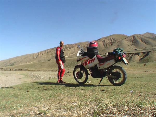 Luggage-laden enduro motorcycle beside a man in red suit, arid hills behind