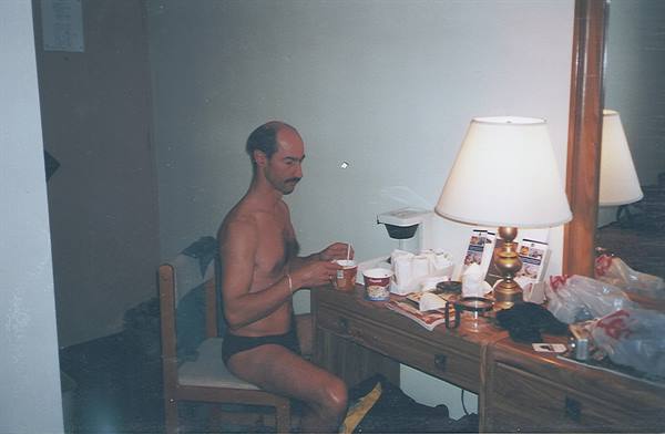 Man in underwear sits at hotel desk, stirring a cup beside the lamp