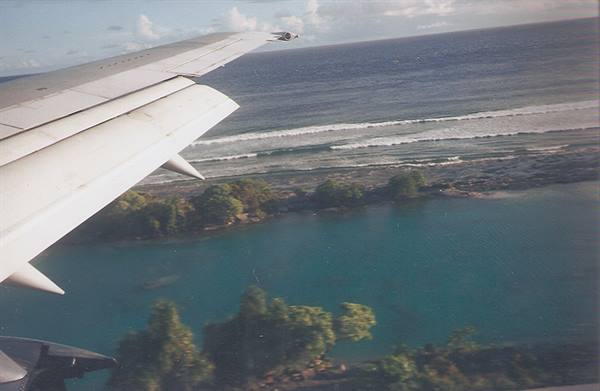 Aircraft wing flying over turquoise lagoon, reef and white breaking waves