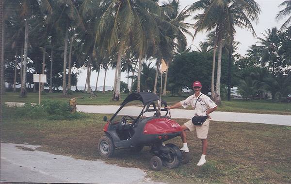 Man in red cap beside a golf cart, palm trees and road behind