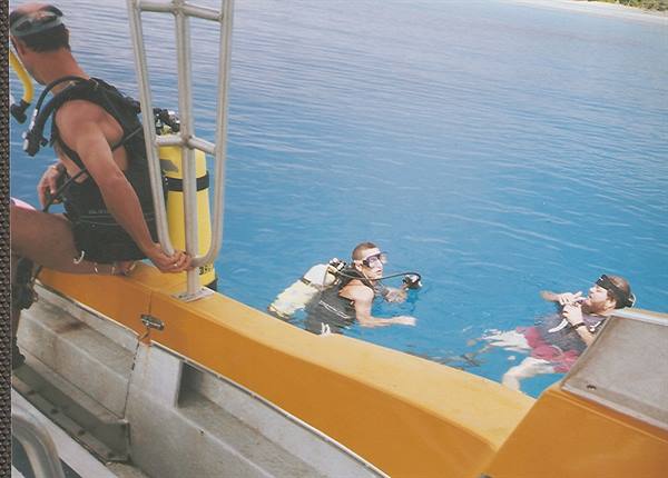 Divers with scuba tanks climb onto a yellow boat in turquoise water