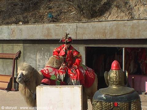 Performer in red imperial costume riding a camel near a warrior statue