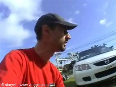 Man in cap and red shirt talking in front of a parked white car