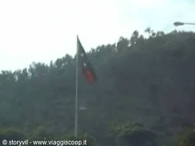 Bangladesh flag waving on a pole, green forested hillside under cloudy sky
