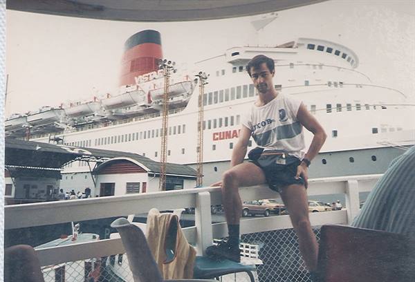 Young man in tank top and shorts sits on a railing beside the Cunard liner
