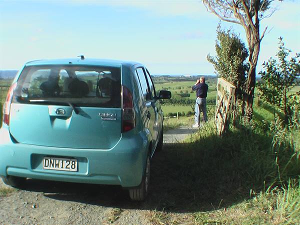 Turquoise car parked on gravel, man photographing green countryside beyond a wooden gate