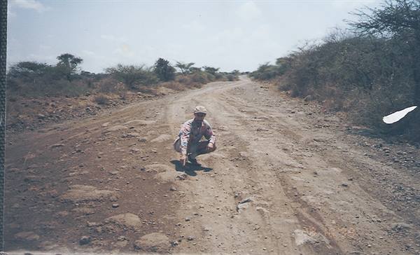Man crouching on rocky dirt road among dry bushes and acacia trees
