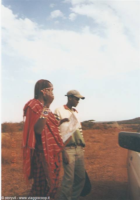Maasai woman with beaded headdress beside a guide in cap near the car