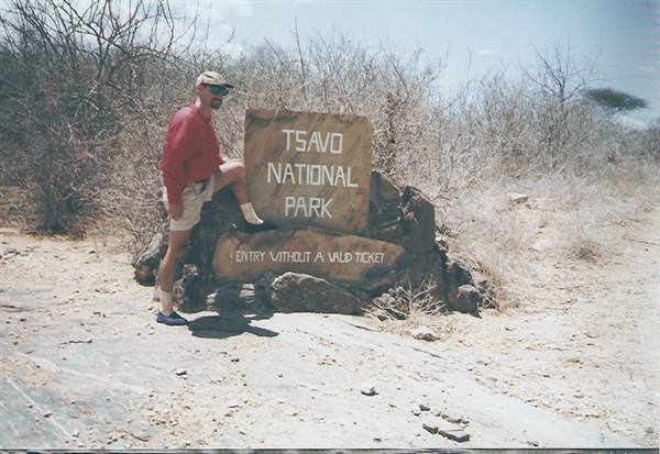 Tourist leaning on the stone sign of Tsavo National Park, dry bushland