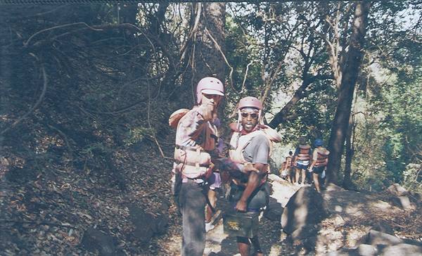 Two men wearing helmets and life jackets walk through woods toward the river