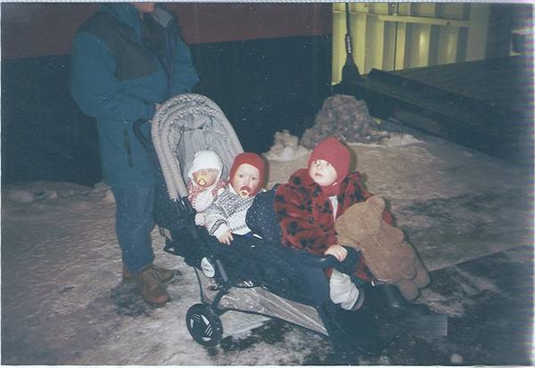 Three children in red hats on a stroller, snowy ground, adult beside