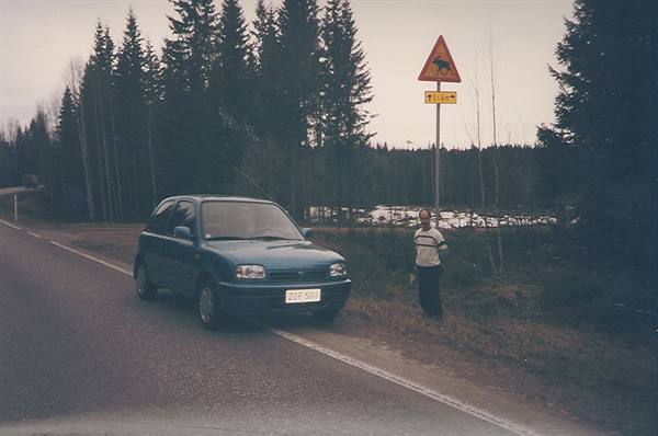 Blue car parked on a country road, man standing beside a moose crossing sign