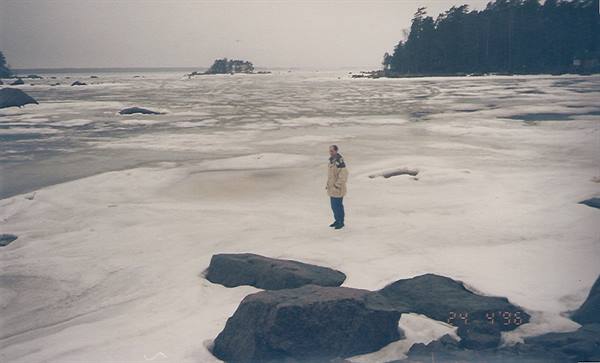 Woman in beige jacket walks across frozen sea, rocks and pines on shore