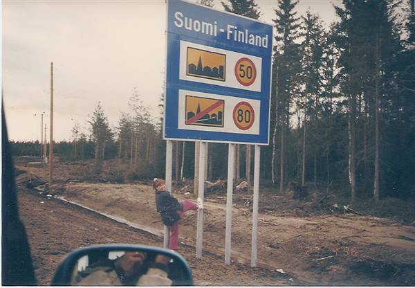 Child leaning on blue Suomi-Finland border road sign, pine forest