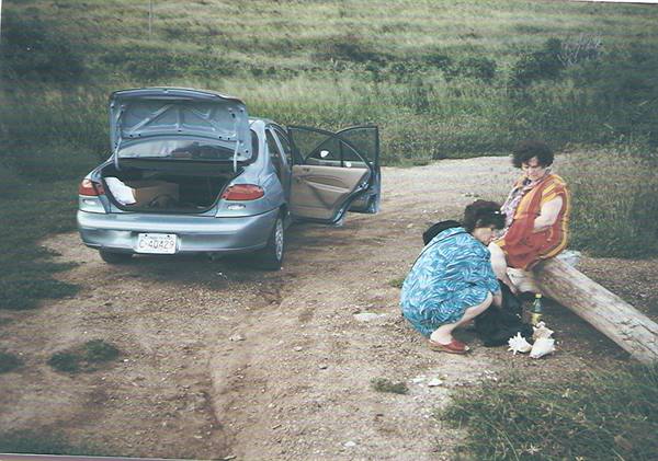 Two women removing shoes by a log, car with open trunk
