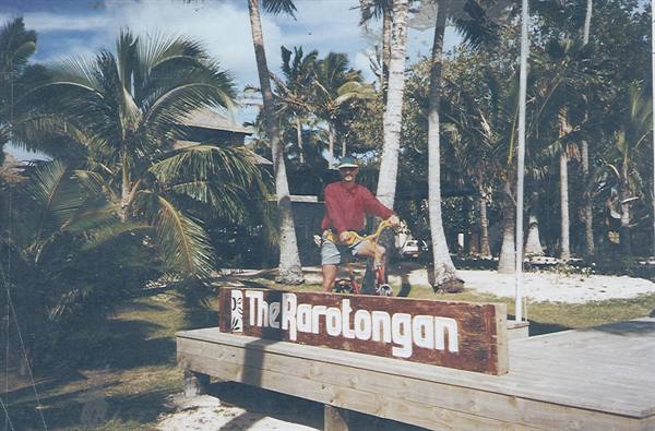 Man in red shirt with bicycle in front of The Rarotongan sign, palm trees