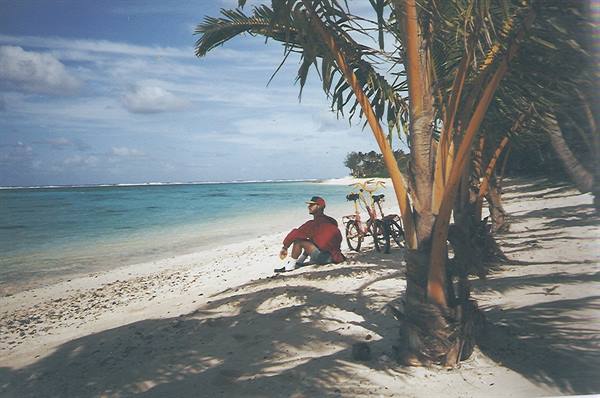 Man in red hat sitting on sand beside two bicycles under palm trees