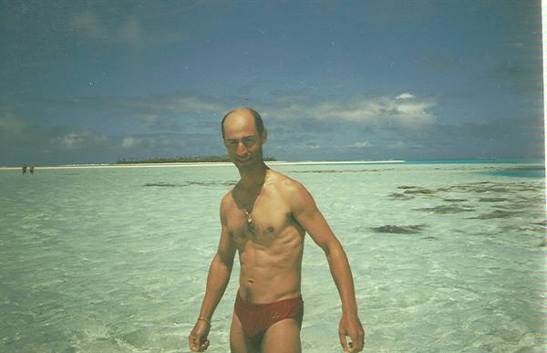 Man in red swim trunks standing in shallow turquoise water, atoll and white beach behind