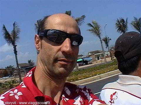 Man in sunglasses and red floral shirt, palm trees and houses behind