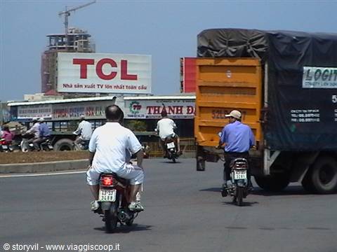 Motorcyclists queuing at the toll booth beside an orange TCL truck