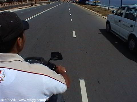 Riding a scooter along a coastal asphalt road, white car nearby
