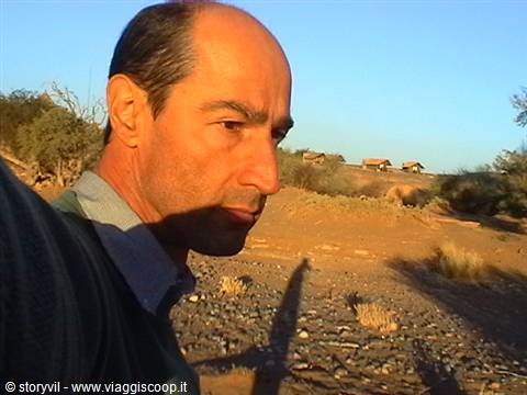 Man close-up on dry rocky terrain, wooden huts in the background