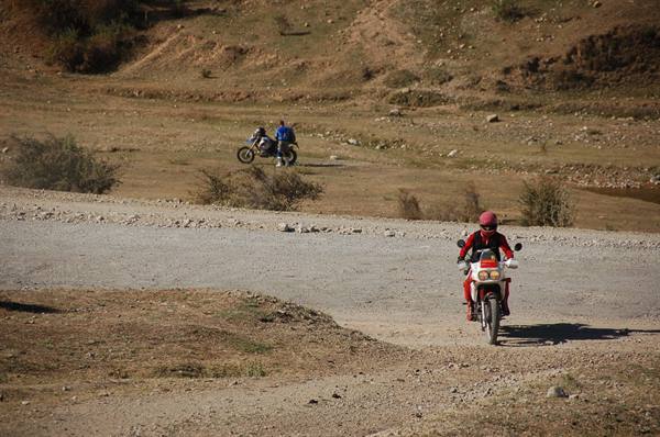Motorcyclist with red helmet rides dirt road, another rider in arid hills