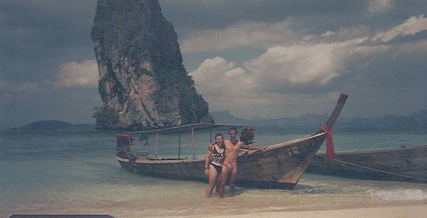 Couple beside wooden longtail boat on beach with limestone cliff, cloudy light