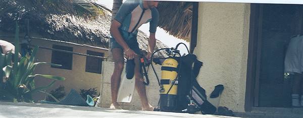 Diver in wetsuit preparing fins and yellow tank under thatched roof