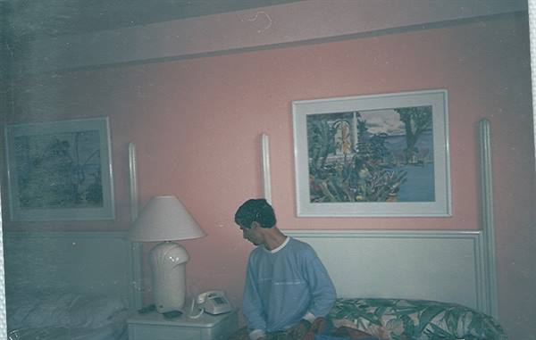 Boy sitting on bed in pink hotel room with lamp and paintings
