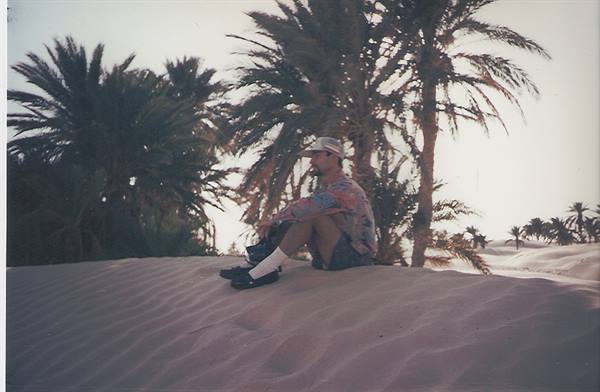 Man sitting on a sand dune beneath palm trees in an oasis