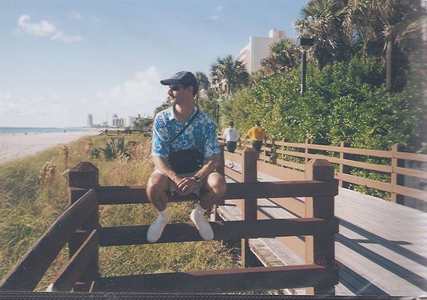 Man in cap and Hawaiian shirt sitting on a wooden fence by a seaside boardwalk