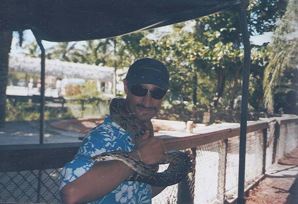 Man in cap and blue shirt holds a python on his shoulders