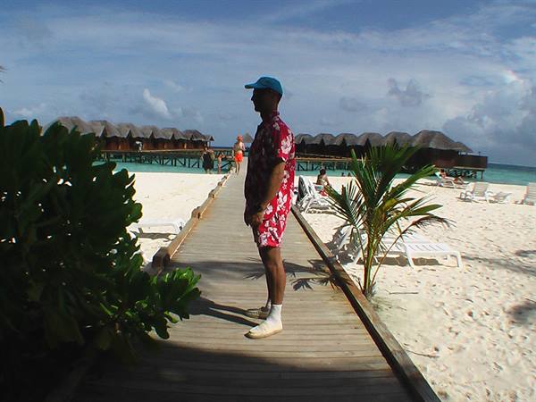 Man in red Hawaiian shirt on wooden boardwalk between sand and overwater bungalows