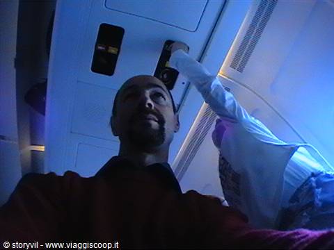 Bearded man looks up at overhead compartments on an airplane under blue reading lights