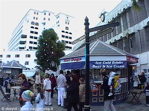 Hot dog kiosk selling cold drinks and beer, people stroll past a signpost
