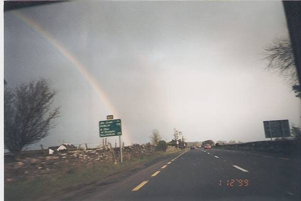 Rainbow over asphalt road, green road sign and stone walls, grey sky